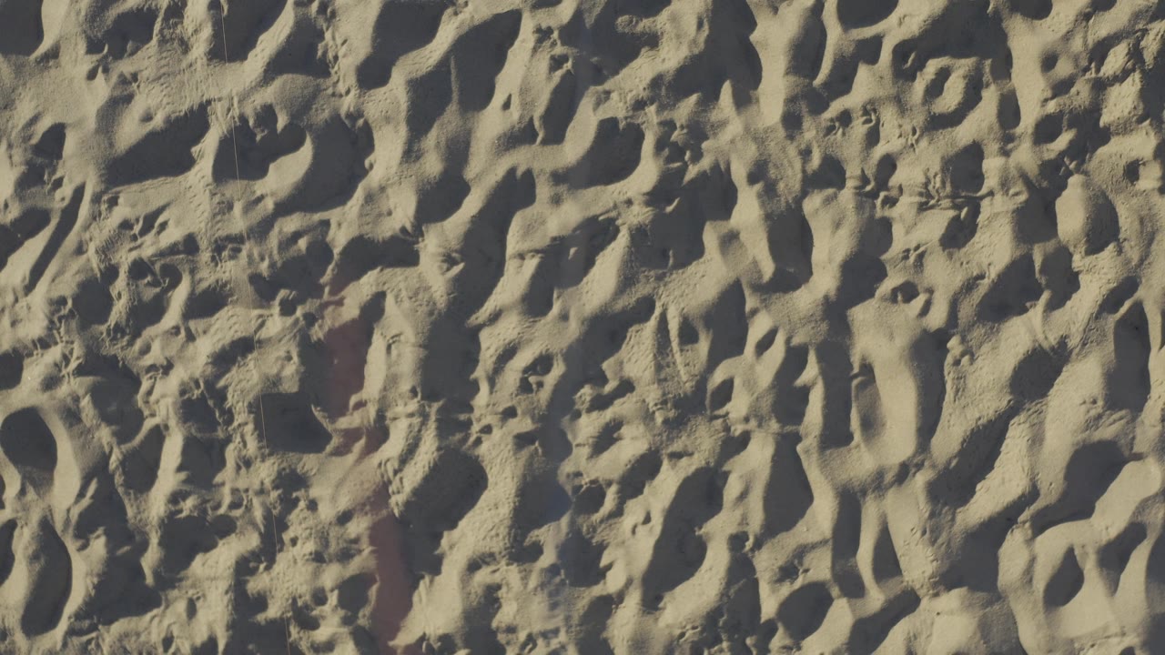 Baja California Sur beach sand fills the frame with a small clear ball near the upper edge, strong sunlight on ridges, dark footprint depressions, and no visible people.