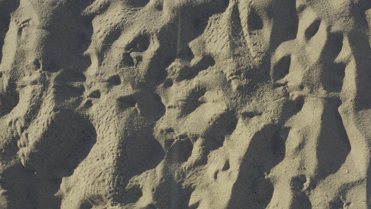 Overhead view of Baja California Sur beach sand with clustered shoe impressions and diagonal shadows emphasizing the uneven surface, with no visible people.