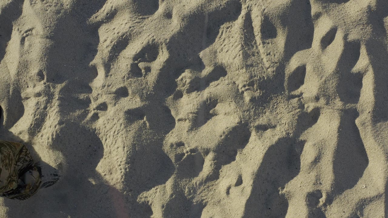 Partial pant leg and shoe at the edge of footprint-marked sand.