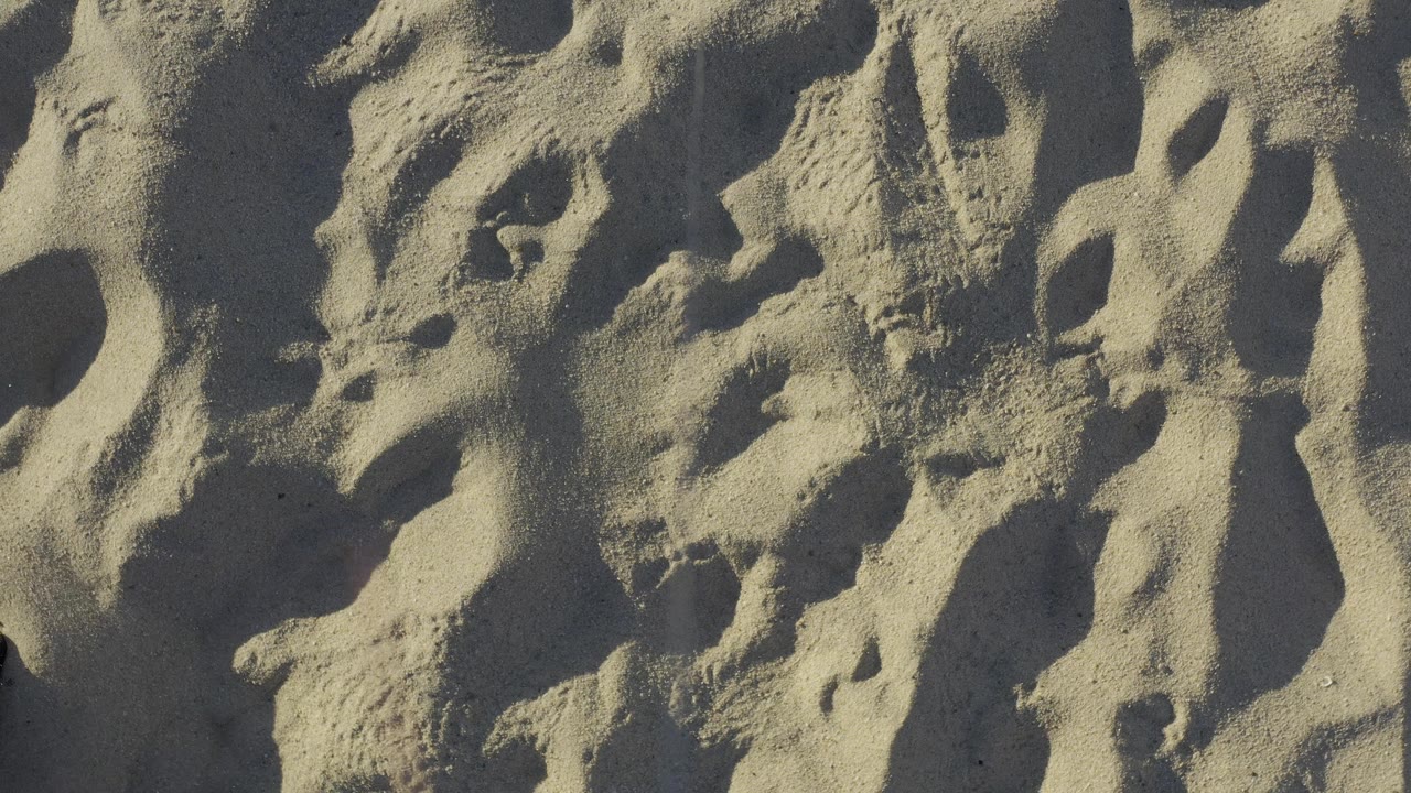 Pale beach sand in Baja California Sur, Mexico, fills the frame with raised ridges, shadowed hollows, and no visible people one second into the clip.