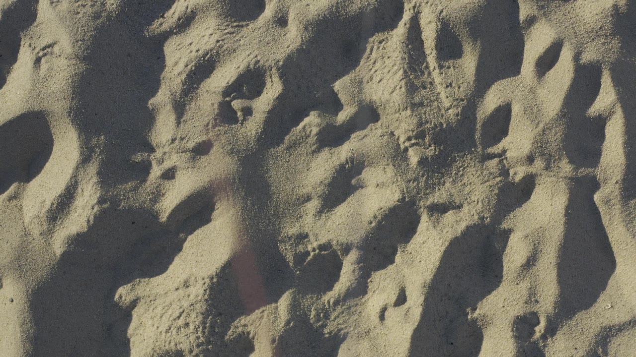 Textured sand with darker pockets and shallow footprint shapes fills the third second in Baja California Sur, Mexico, with no visible people.