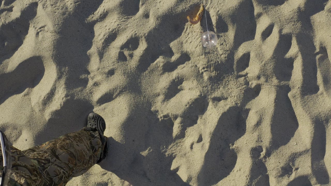 Close downward view of rippled Baja California Sur beach sand with a partial boot and pant leg at the lower left, a small dry leaf or shell near the top, and no identifiable people.