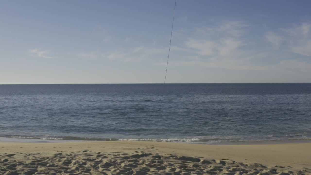 Wide coastal view in Baja California Sur with textured sand foreground, calm blue sea, a straight horizon, faint high clouds, and no visible people at 0:02.