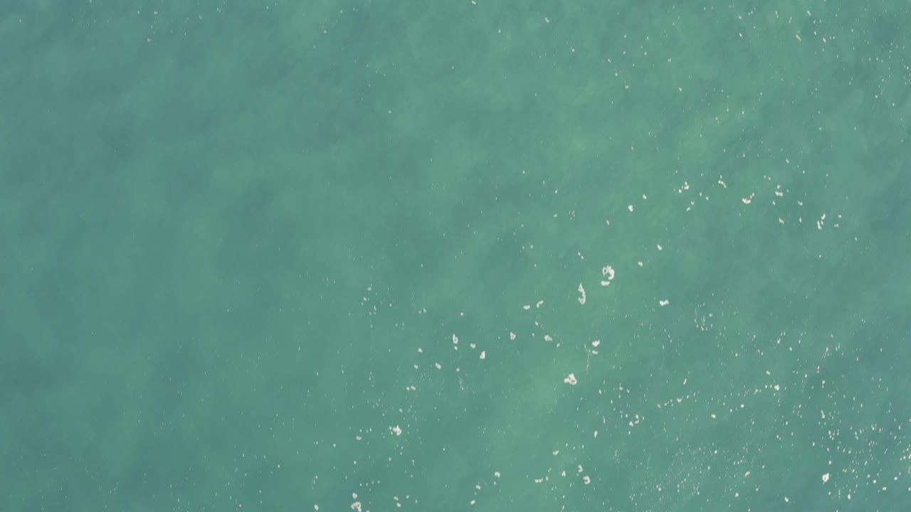 Top-down aerial water view in Baja California Sur, Mexico, with faint darker underwater patches visible through the turquoise sea and no visible people.