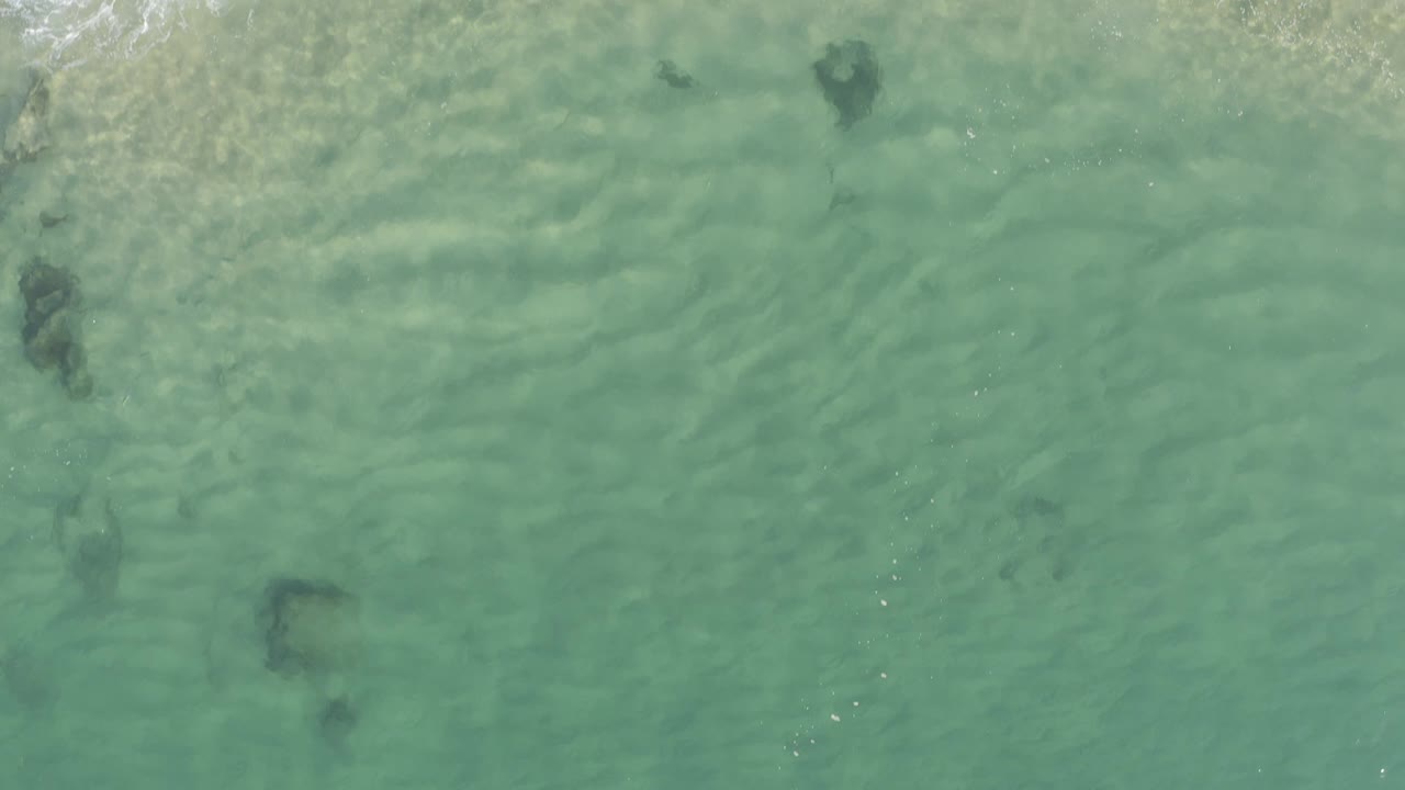 Top-down aerial view of turquoise water in Baja California Sur, Mexico, with dark submerged rock or reef patches and no visible people.