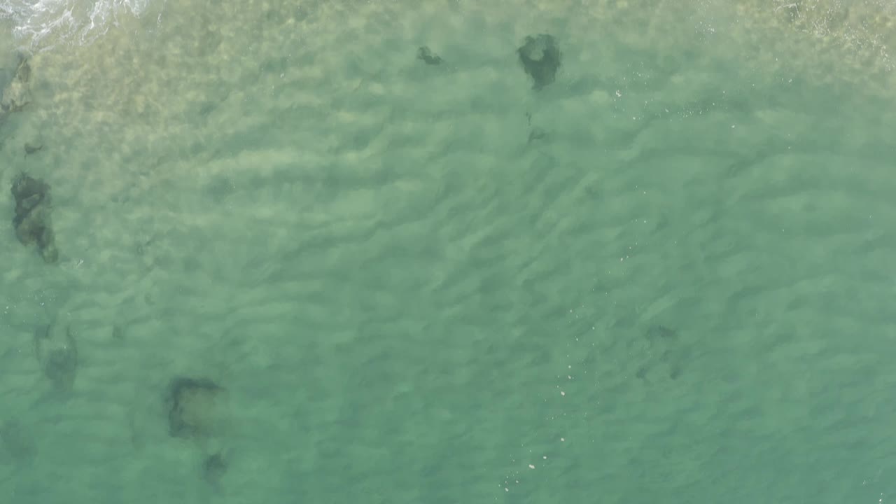 Aerial frame over clear green-blue shallows in Baja California Sur, Mexico, with scattered underwater dark shapes and no visible people.