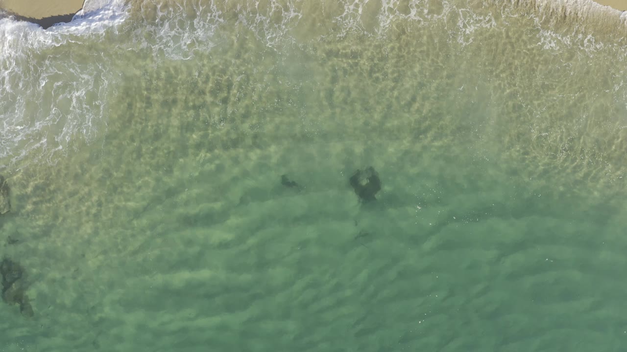 Overhead Baja California Sur shoreline view with white surf entering turquoise shallows, a small strip of sand, and no visible people.