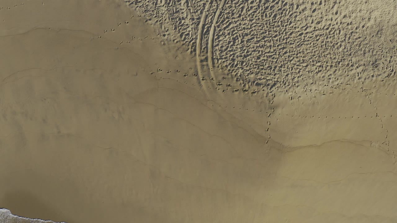 Top-down aerial view of Baja California Sur beach sand with curved tire tracks, rippled dry sand, smoother flat patches, and no visible people.