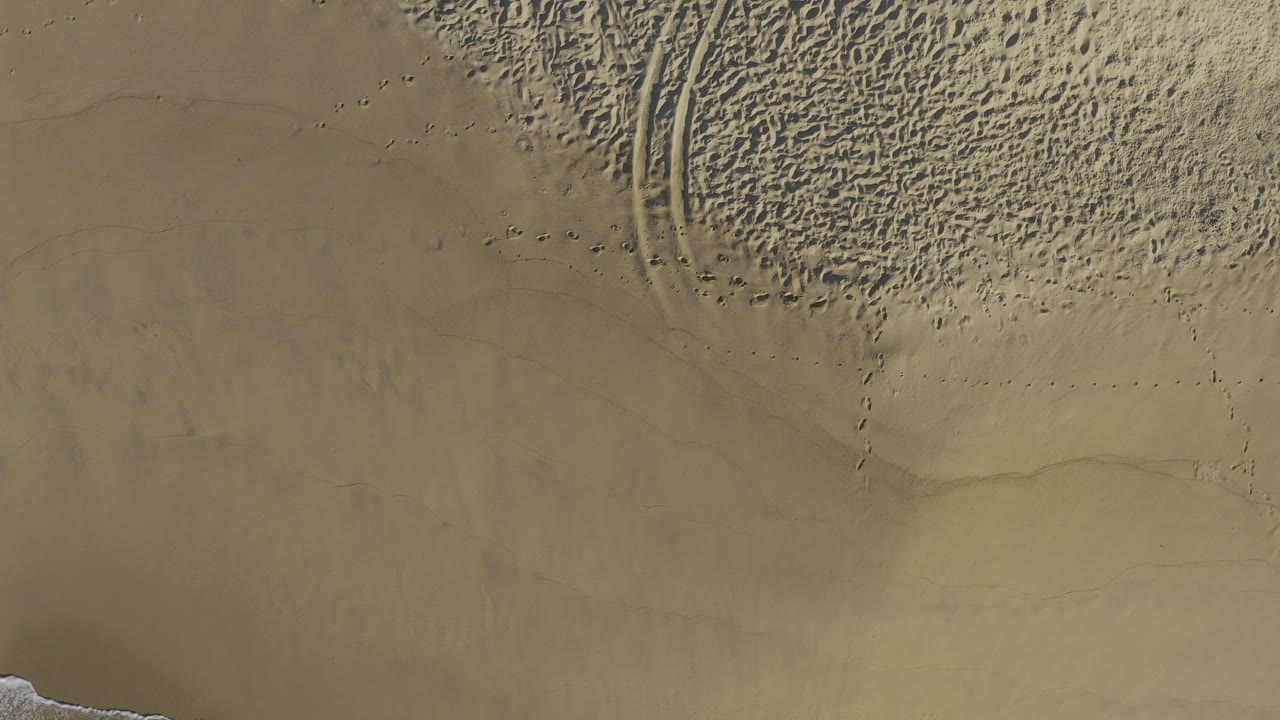 Overhead sand textures with tire marks crossing the beach.