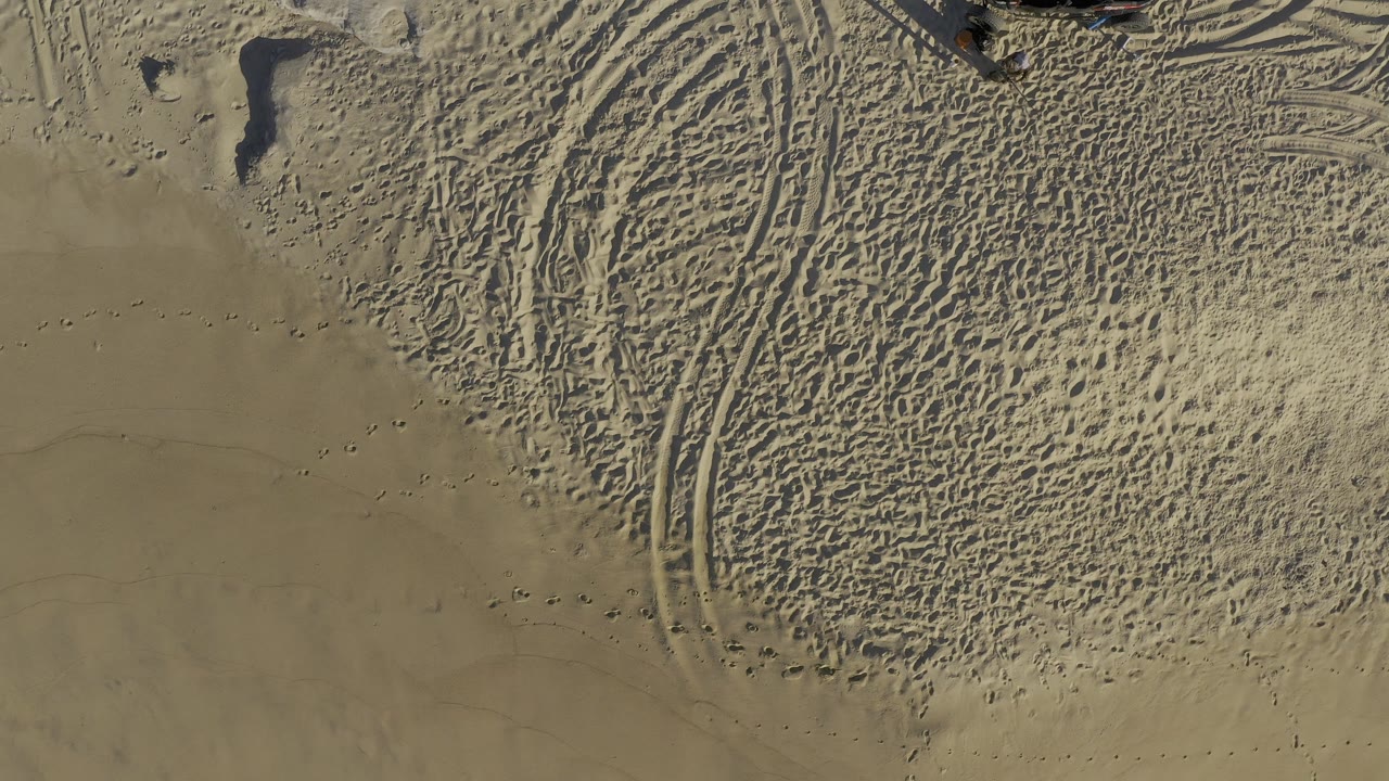 Overhead view of Baja California Sur sand with curved tire tracks, scattered footprints, dimpled texture, and no visible people at the opening frame.