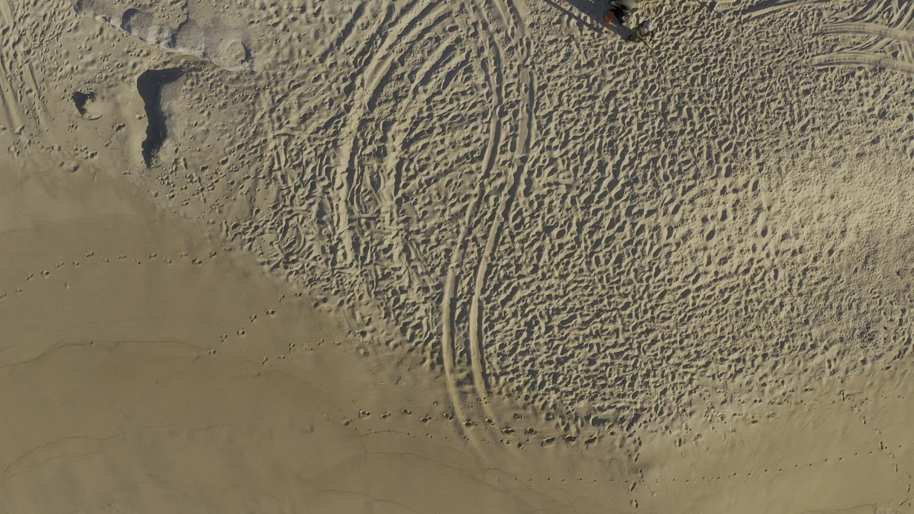 Overhead view of pale sand in Baja California Sur, Mexico, with curved tire tracks, scattered footprints, small dark rocks, and no visible people in this frame.