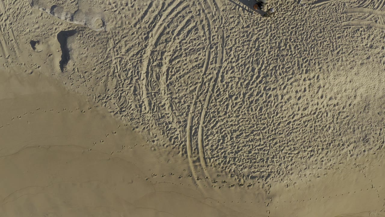Top-down Baja California Sur beach sand shows dense footprints, arcing tire marks, and small rock shadows, with no visible people in this moment.
