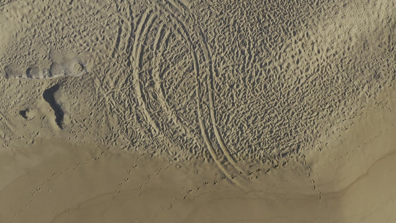 Aerial beach view in Baja California Sur, Mexico, with rippled sand, footprints, a pair of tire tracks along the right side, and no visible people in the frame.
