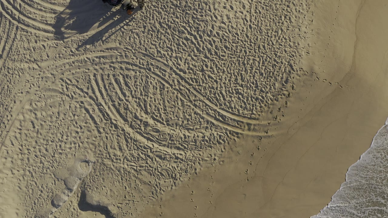 Overhead sand with tire tracks and a small dark object at upper left.