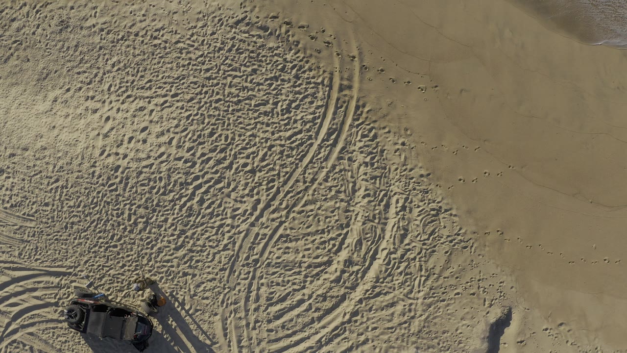 Aerial view in Baja California Sur, Mexico, showing tire ruts, footprints, textured tan sand, and the corner of a dark vehicle at the lower left with no visible people.