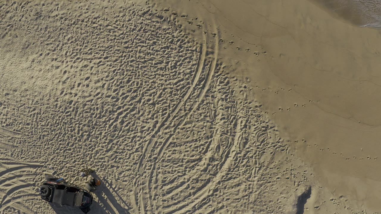 Aerial frame from Baja California Sur, Mexico, with curving tire tracks crossing textured sand and a dark vehicle partly visible near the lower edge, with no visible people.