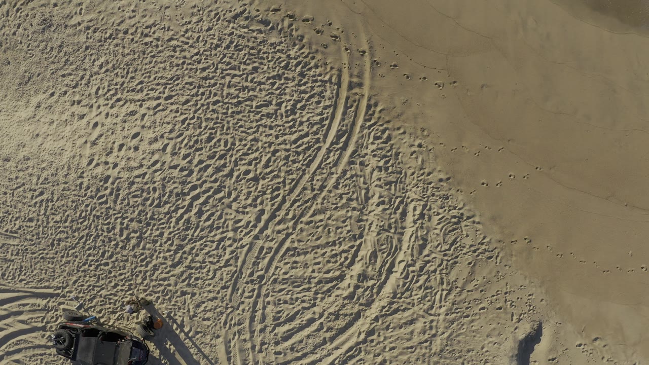 Overhead Baja California Sur sand scene with dense tire ruts, scattered footprints, smoother sand to the upper right, and a partial dark vehicle at the bottom with no visible people.