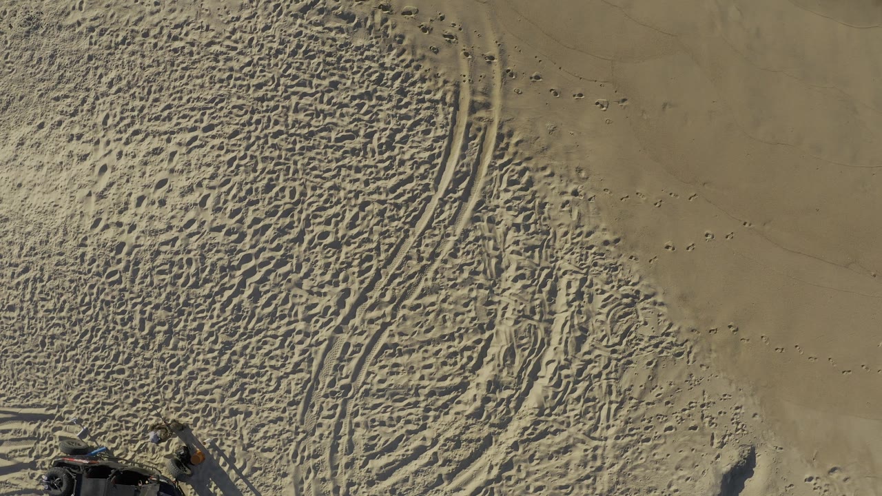 Aerial view over Baja California Sur coastal sand, showing parallel tire tracks, many footprints, and the corner of a dark off-road vehicle at the lower left, with no visible people.