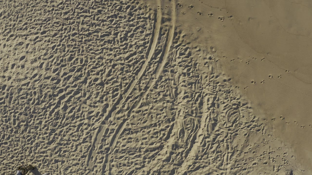 Overhead Baja California Sur, Mexico frame where diagonal grooves run through rippled tan ground, with scattered small impressions across the smoother upper area and no visible people.