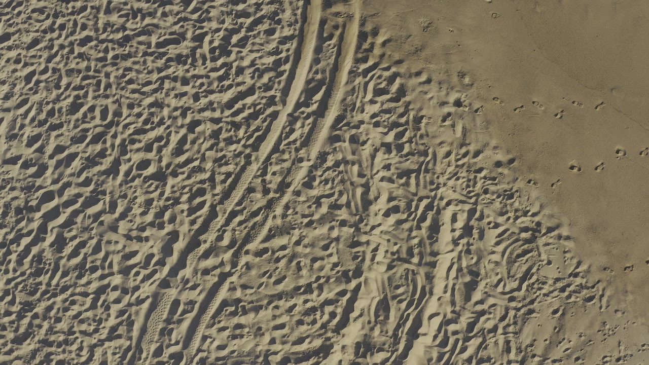 Aerial view of Baja California Sur beach sand where two tire-track lines run near the left edge and footprints cluster across the center, with no visible people.