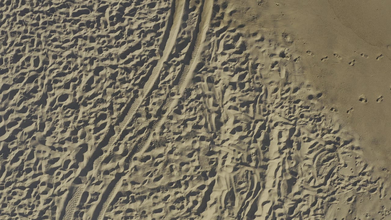 Overhead Baja California Sur sand view with a pronounced curved tire path at left, dense footmarks through the center, smoother tan sand at upper right, and no visible people.