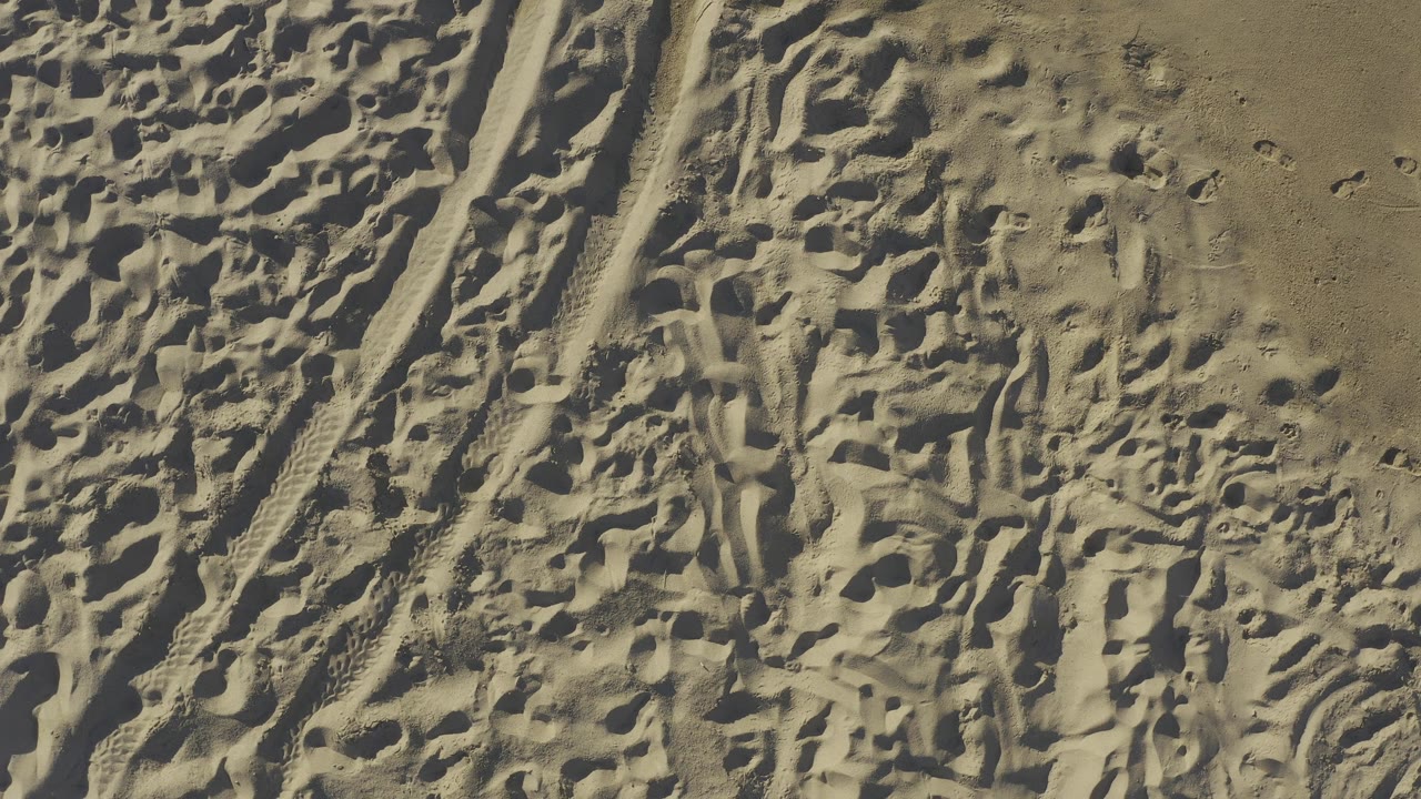 High-angle view of textured beach sand in Baja California Sur, Mexico, showing irregular footprints, tire-like grooves, and no visible people.