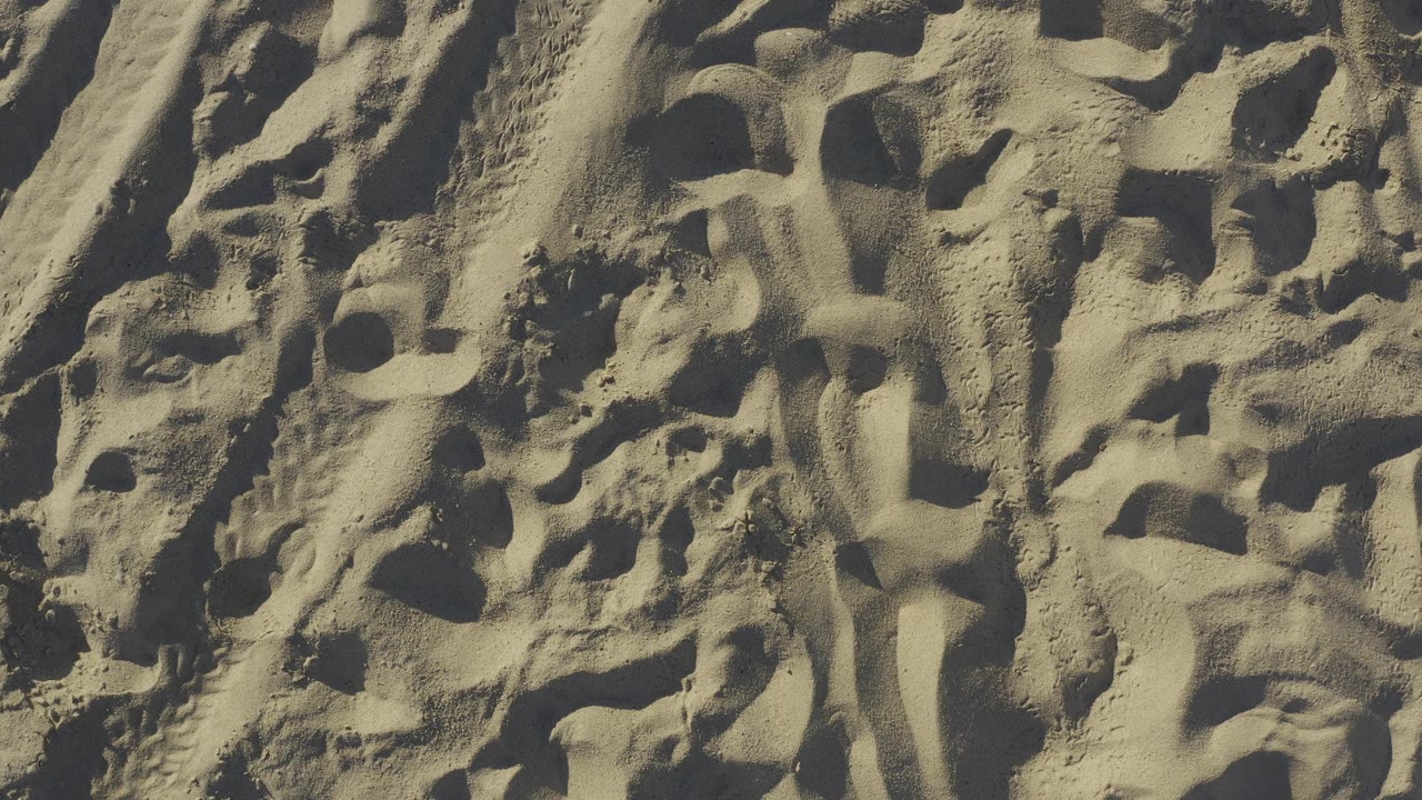 Sunlit overhead beach sand in Baja California Sur, Mexico, with raised mounds, deeper dark marks, fine grain detail, and no visible people.