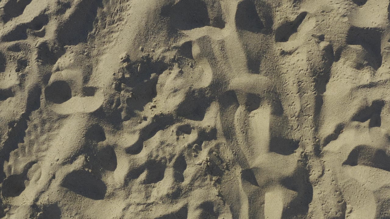 Tight overhead view of Baja California Sur beach sand with elongated ridges, hollowed marks, warm beige color, and no visible people.
