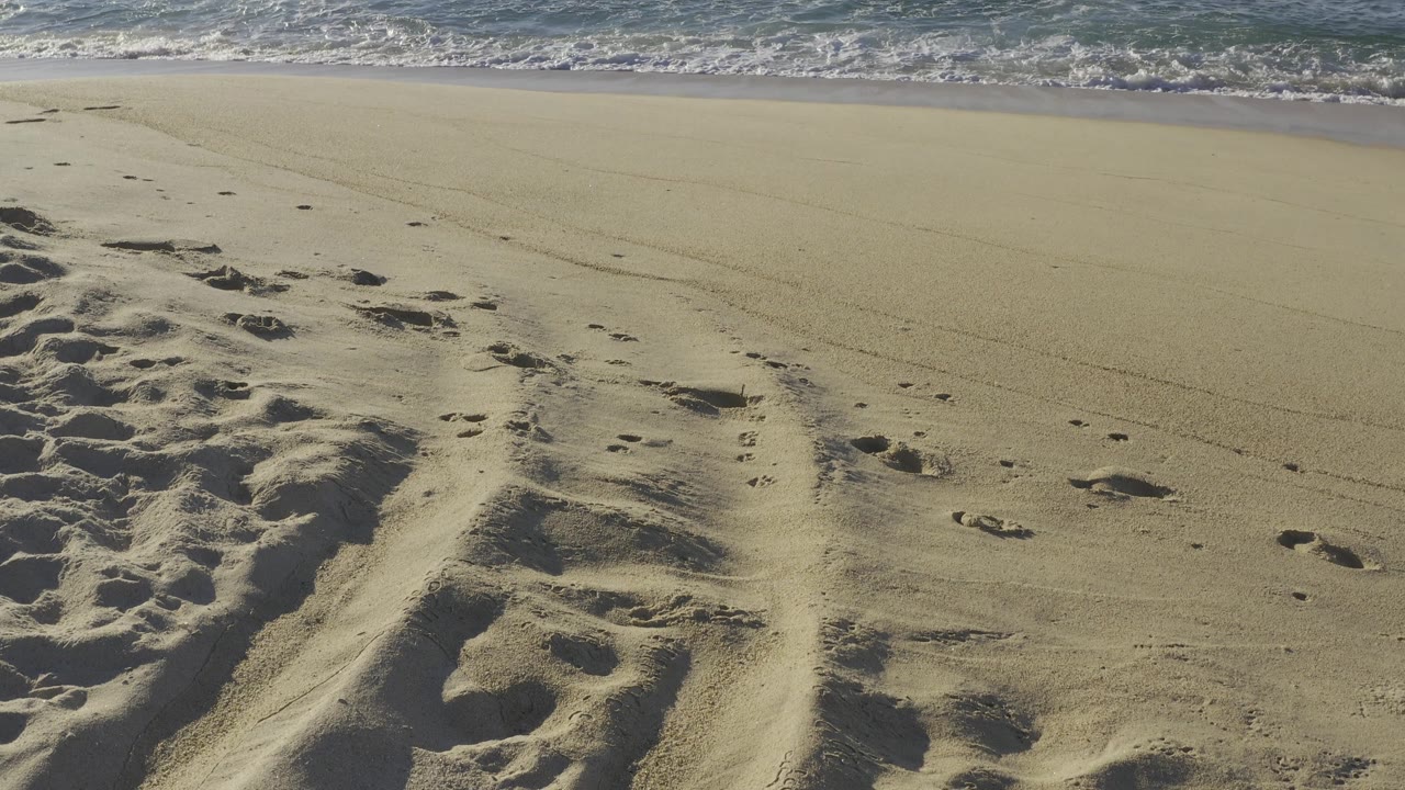 Low shoreline view in Baja California Sur, Mexico, with footprint tracks across tan sand and a strip of foamy surf near the top of frame, no visible people.