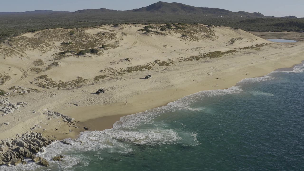 Aerial frame over Baja California Sur, Mexico showing teal surf in the foreground, rocks at lower left, pale beach and dunes beyond, and tiny people on the sand.