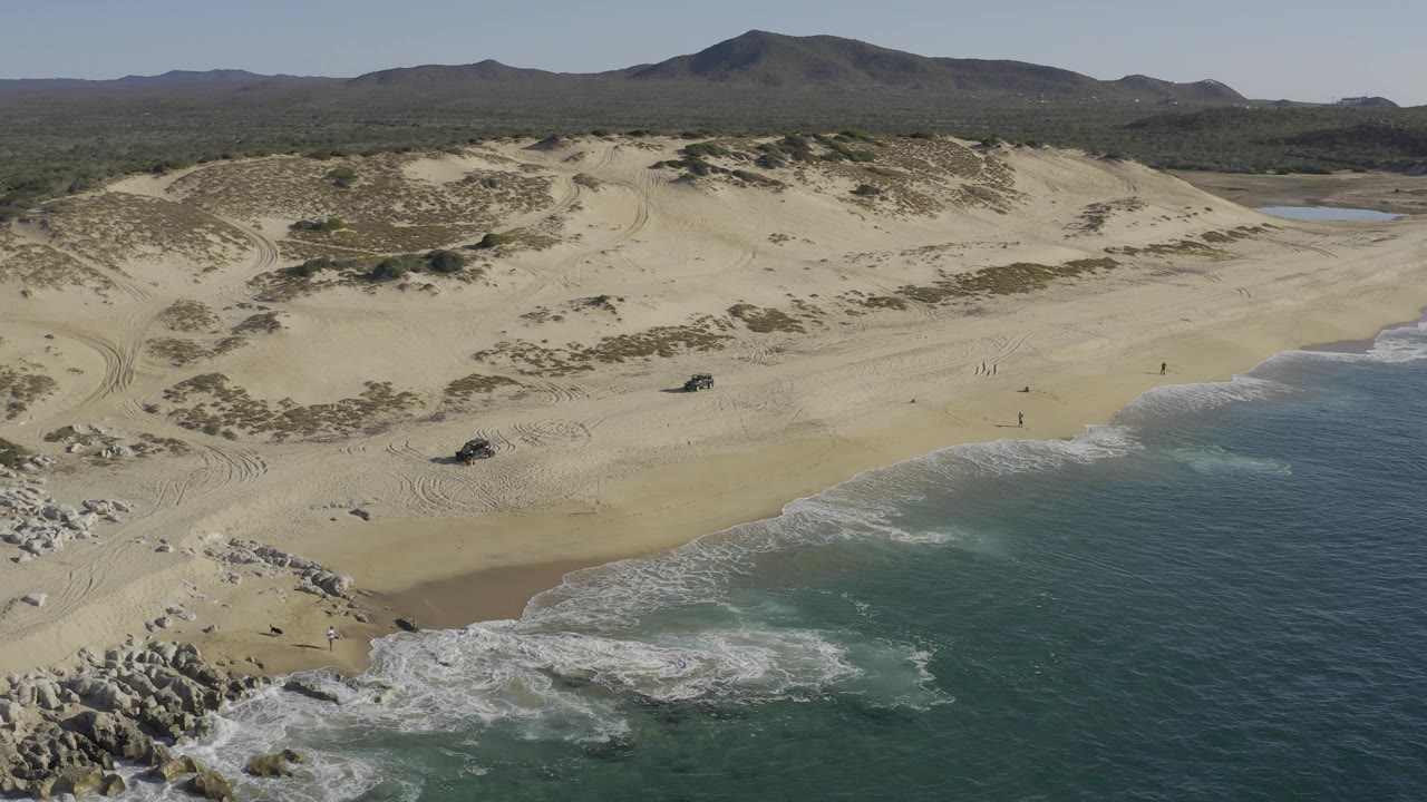 Baja California Sur aerial frame showing more of the beach curve at right, pale dunes across the center, dark scrubland behind them, and tiny people scattered on the sand.