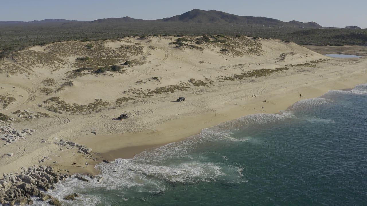 Wide aerial coastline with dunes, rocky foreground, surf, and mountains inland.