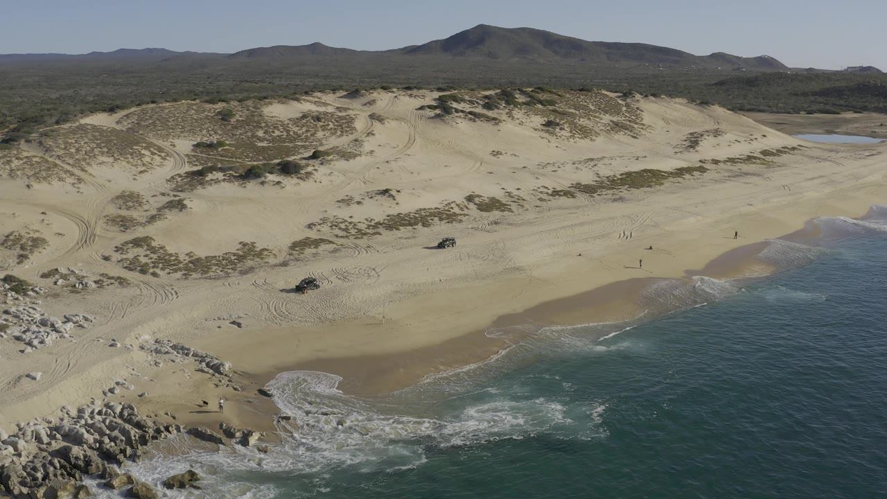 Drone frame over Baja California Sur coastline with the waterline shifted farther right, showing a broad sand slope, surf foam, parked off-road vehicles, tiny people, scrub, and mountains.