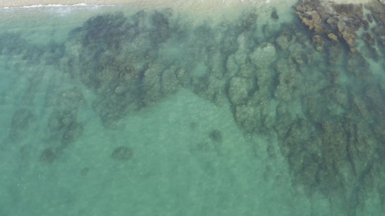 Aerial Baja California Sur shoreline frame with submerged rock clusters filling the water, a narrow white surf line near the top edge, and no visible people.