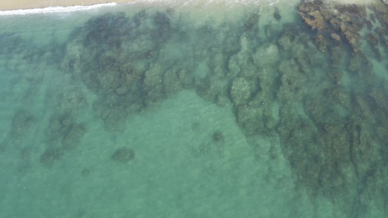 Top-down view in Baja California Sur, Mexico, with broad dark rock formations under pale water and the sandy beach barely visible along the top edge, no visible people.