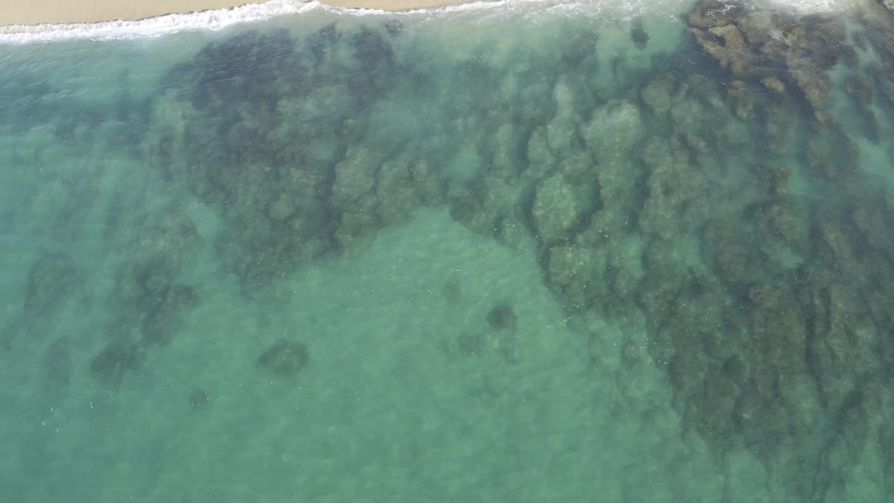 Aerial frame from the Baja California Sur coast showing tan rocks beneath blue-green water, with white foam tracing the upper shoreline and no visible people.