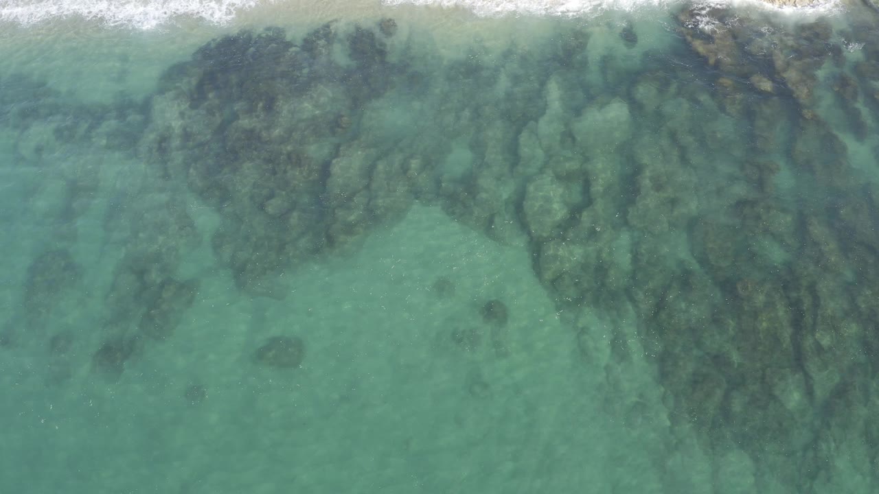 High overhead view in Baja California Sur, Mexico, showing turquoise shallow water, dark submerged rock formations, and a narrow band of breaking surf along the top edge, with no visible people.