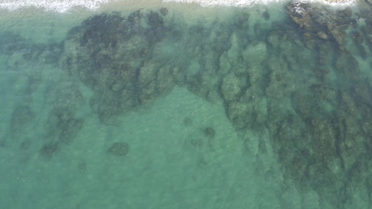 Clear green-blue water over dark underwater rock patterns near breaking waves.