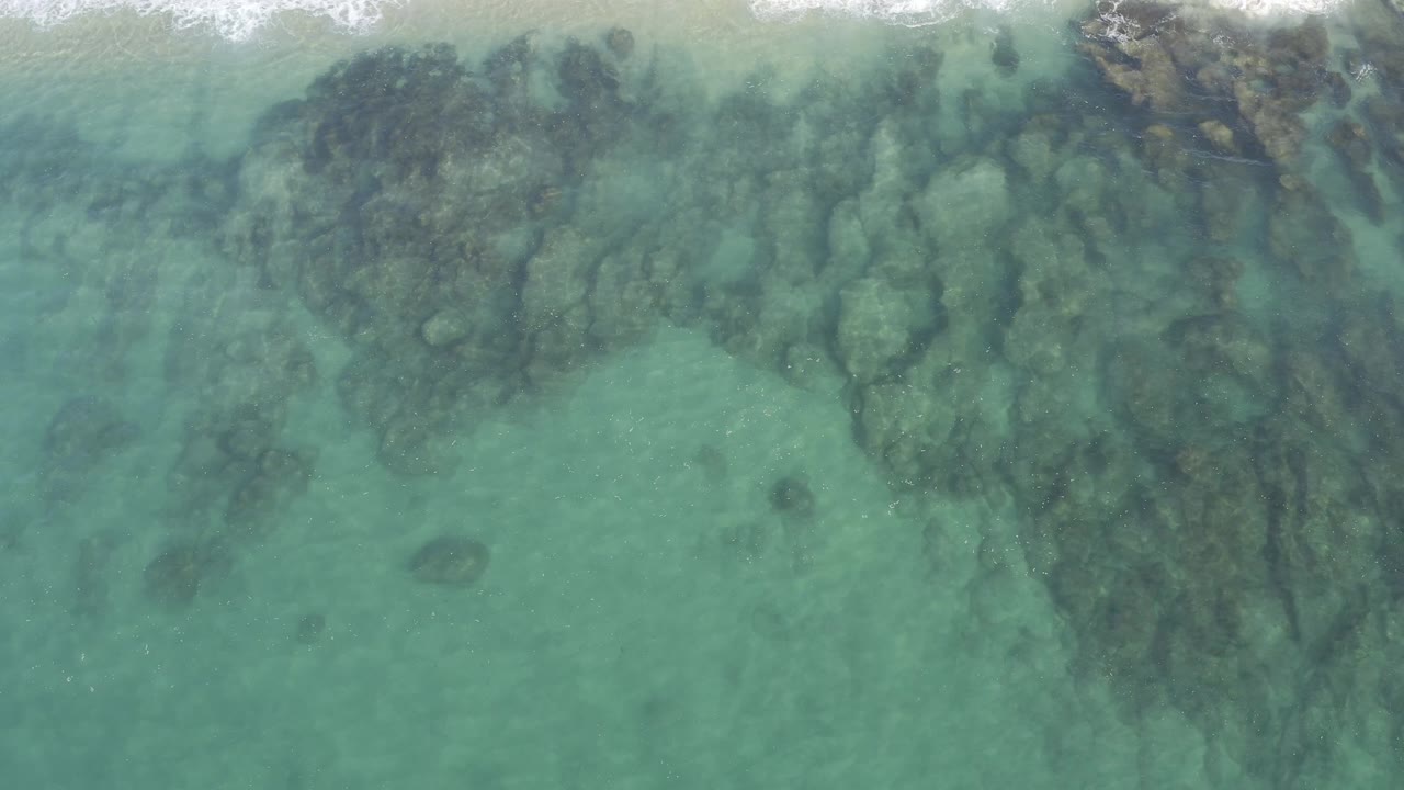 Top-down view in Baja California Sur, Mexico, where reef-like dark shapes spread under turquoise water and a thin surf band breaks across the upper frame, with no visible people.