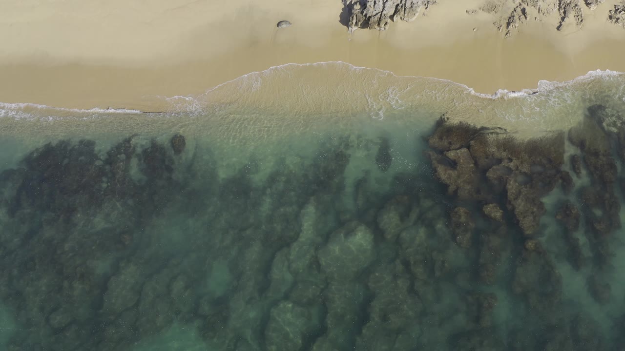 Overhead Baja California Sur shoreline with a broader patch of white foam on the beach edge, clear water below, and dark submerged rock formations, with no visible people.