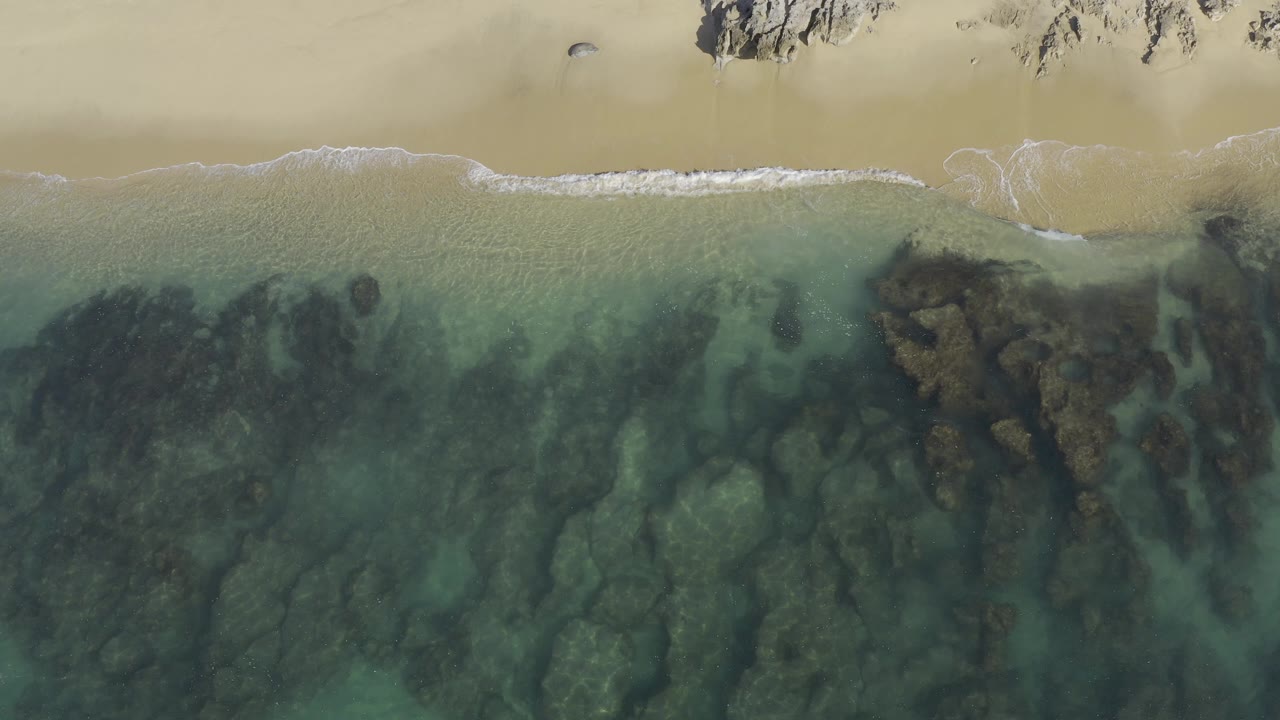 Overhead Baja California Sur beach view with transparent green-blue water over dark rocks, pale sand along the top edge, a narrow white wave line, and no visible people.
