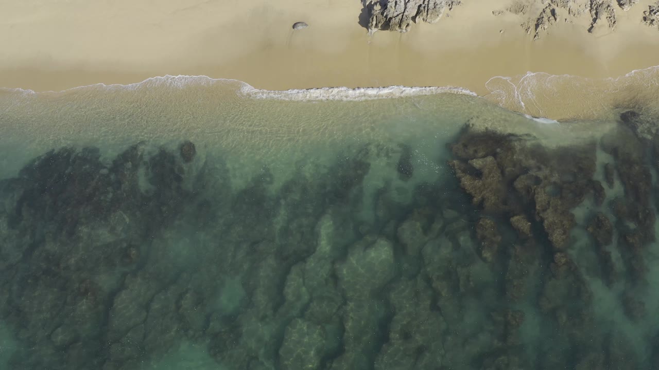 Aerial view in Baja California Sur with shallow transparent water crossing dark rock shapes, smooth pale sand at the top, faint foam at the shore, and no visible people.