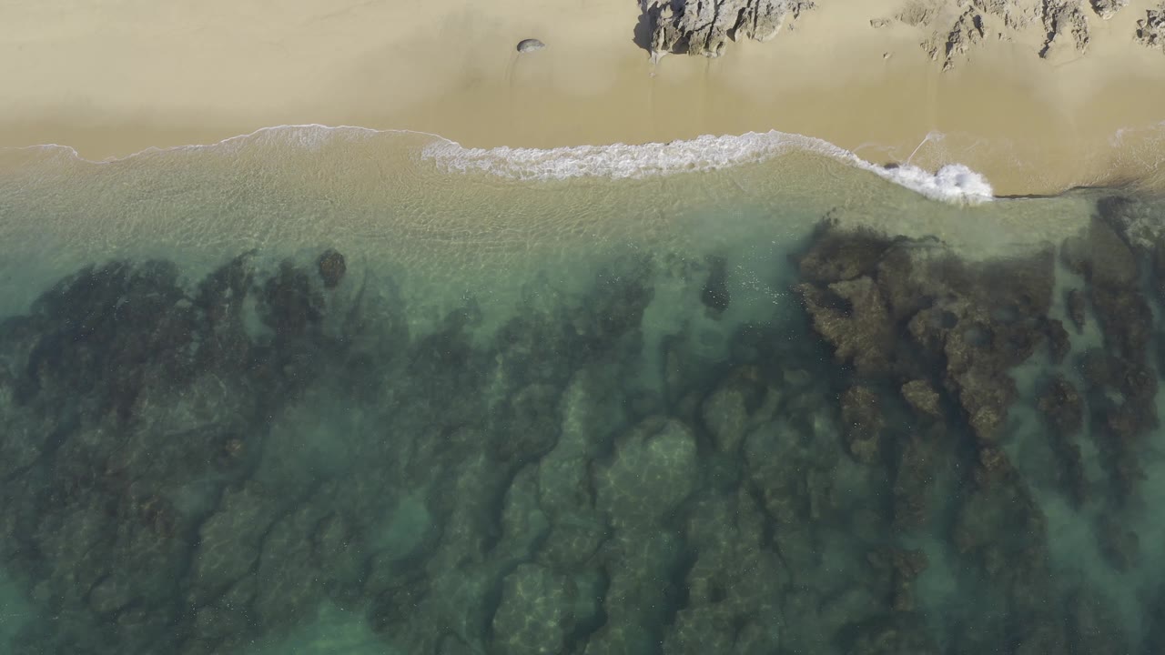 Top-down Baja California Sur shoreline frame showing a brighter white wave breaking near the sand, transparent water below, dark rock patches, and no visible people.