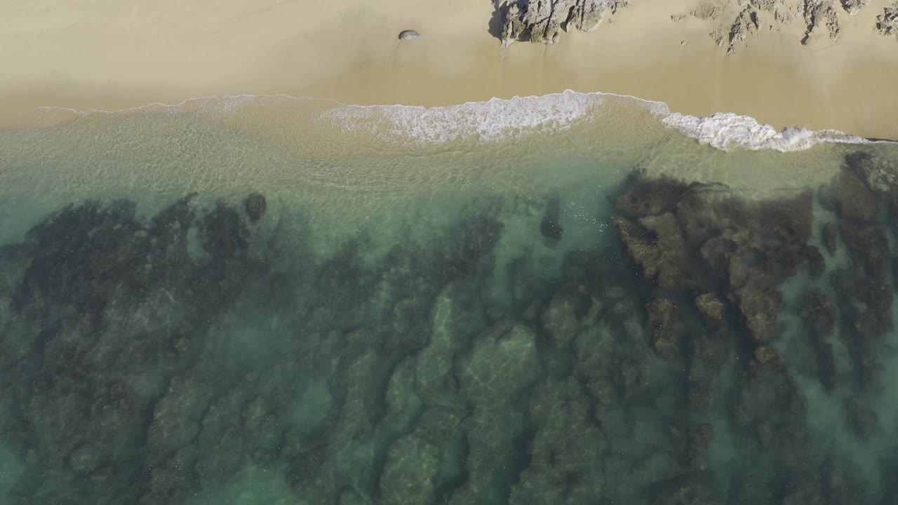 Aerial shoreline with a foamy wave curling across shallow water and rocks.