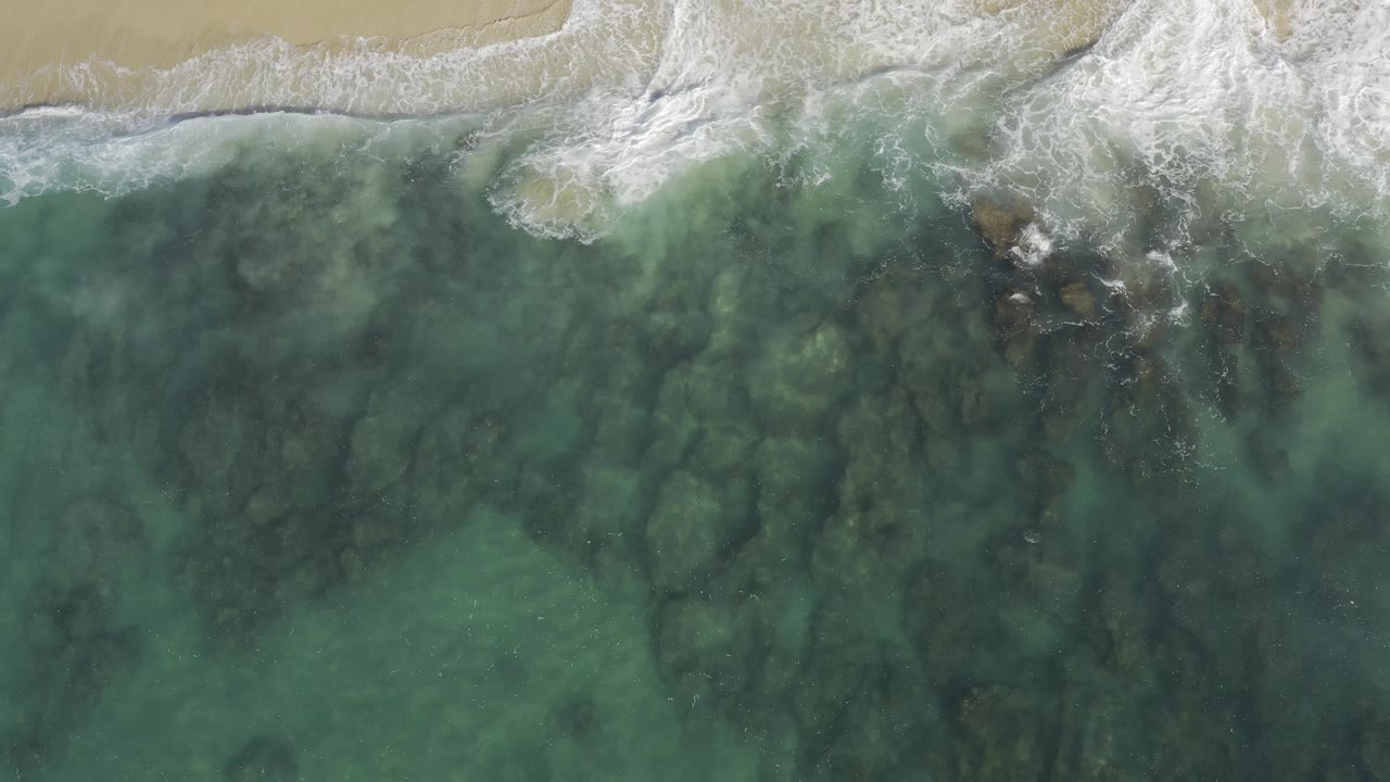 Overhead view of white surf washing onto pale sand above clear green-blue water in Baja California Sur, Mexico, with no visible people.
