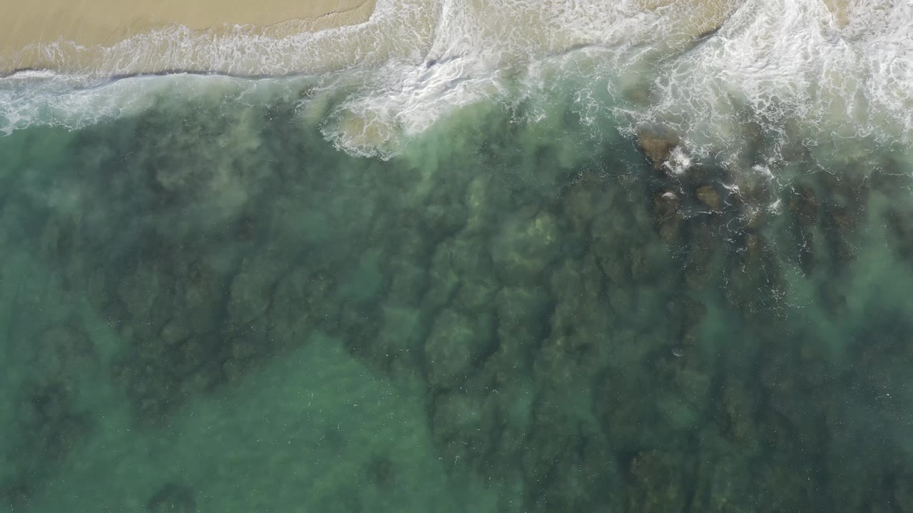 Top-down Baja California Sur shoreline frame with white foam spreading along the upper beach edge and darker underwater forms below, with no visible people.