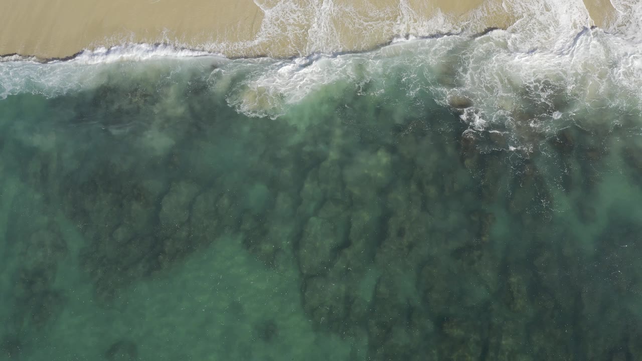 Overhead coastal view in Baja California Sur, Mexico, where a breaking wave crosses pale sand and green water reveals dark reef-like texture below, with no visible people.