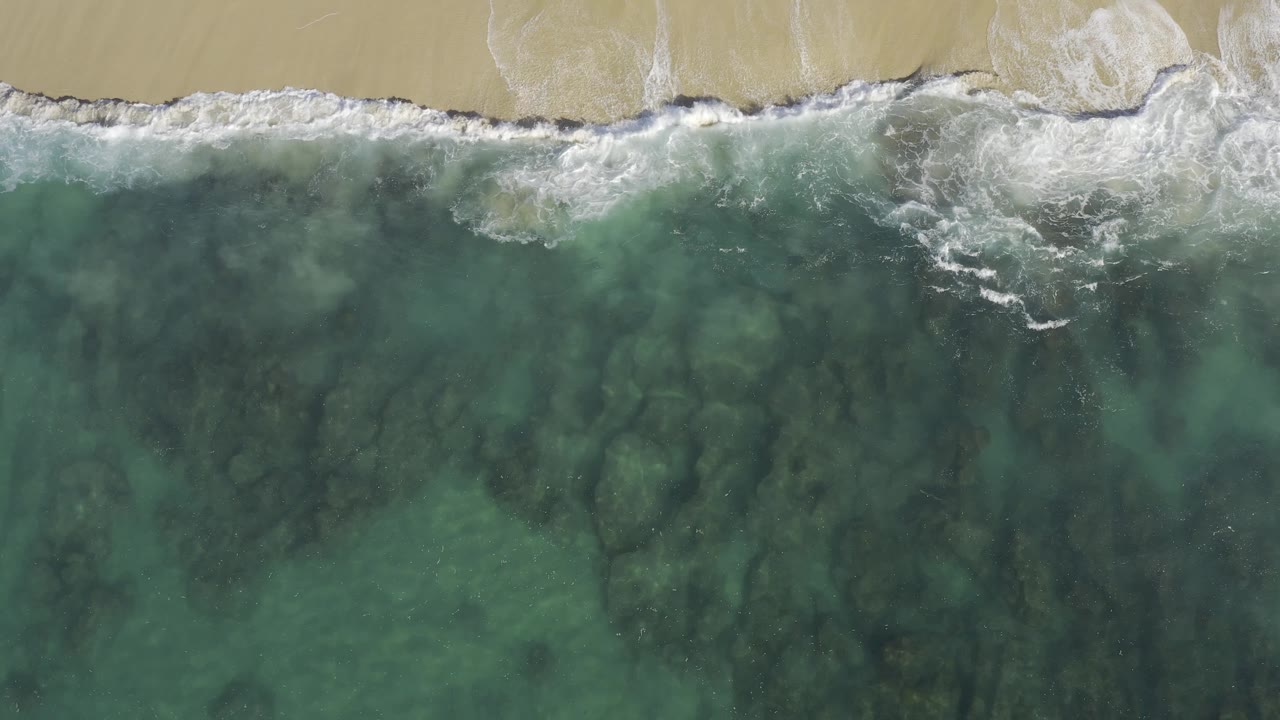 Aerial view of a longer foamy surf band running across the top of clear green-blue water on the Baja California Sur coast, with no visible people.