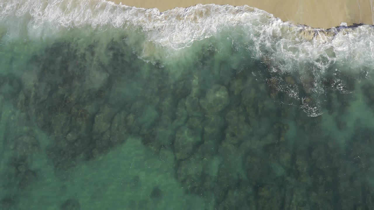 Overhead view in Baja California Sur, Mexico with white surf crossing the top edge, green shallow water below, dark submerged rock patterns, and no visible people.