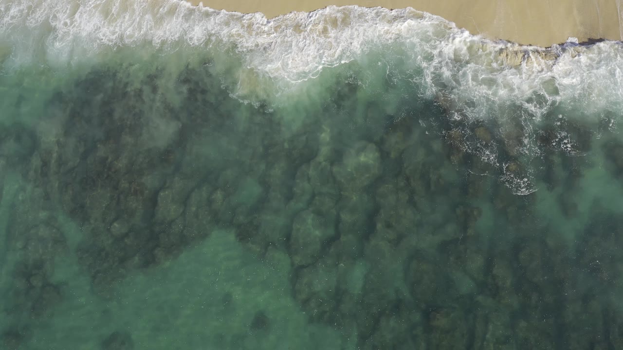 Aerial shoreline frame in Baja California Sur, Mexico with foamy water near the upper edge and darker reef-like rock shapes visible under the green sea, with no visible people.